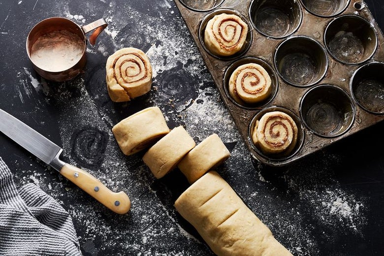 A log of raw cinnamon rolls. Some have been sliced and put into the cups of a muffin tin.